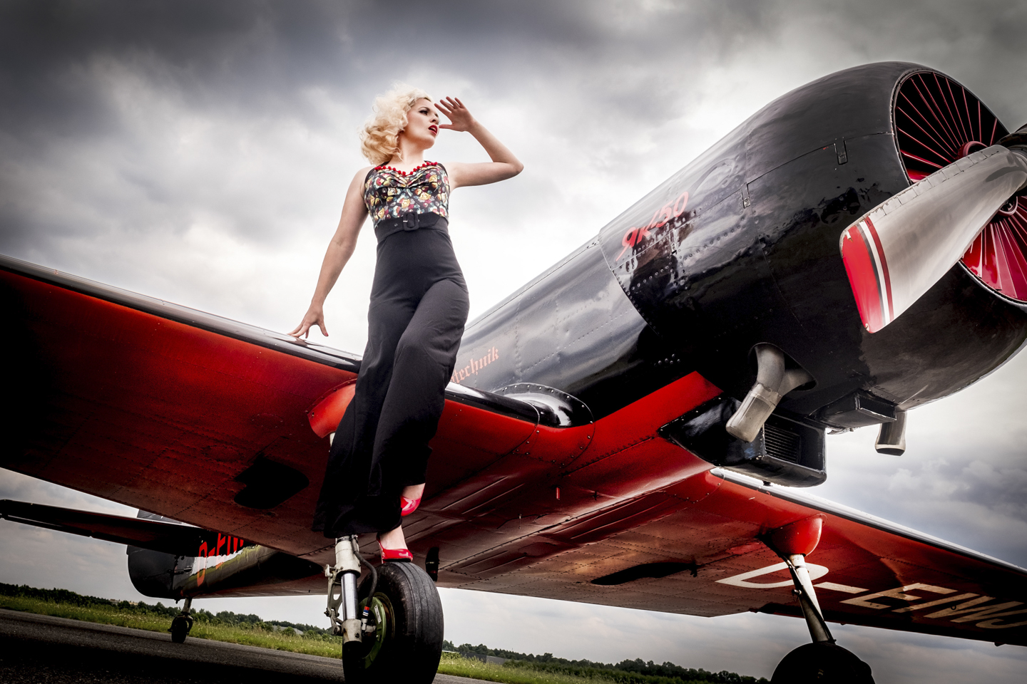Blonde Frau im 50er Jahre Hosenanzug steht auf dem Reifen eines kleinen Propeller Flugzeugs und schaut in die Ferne