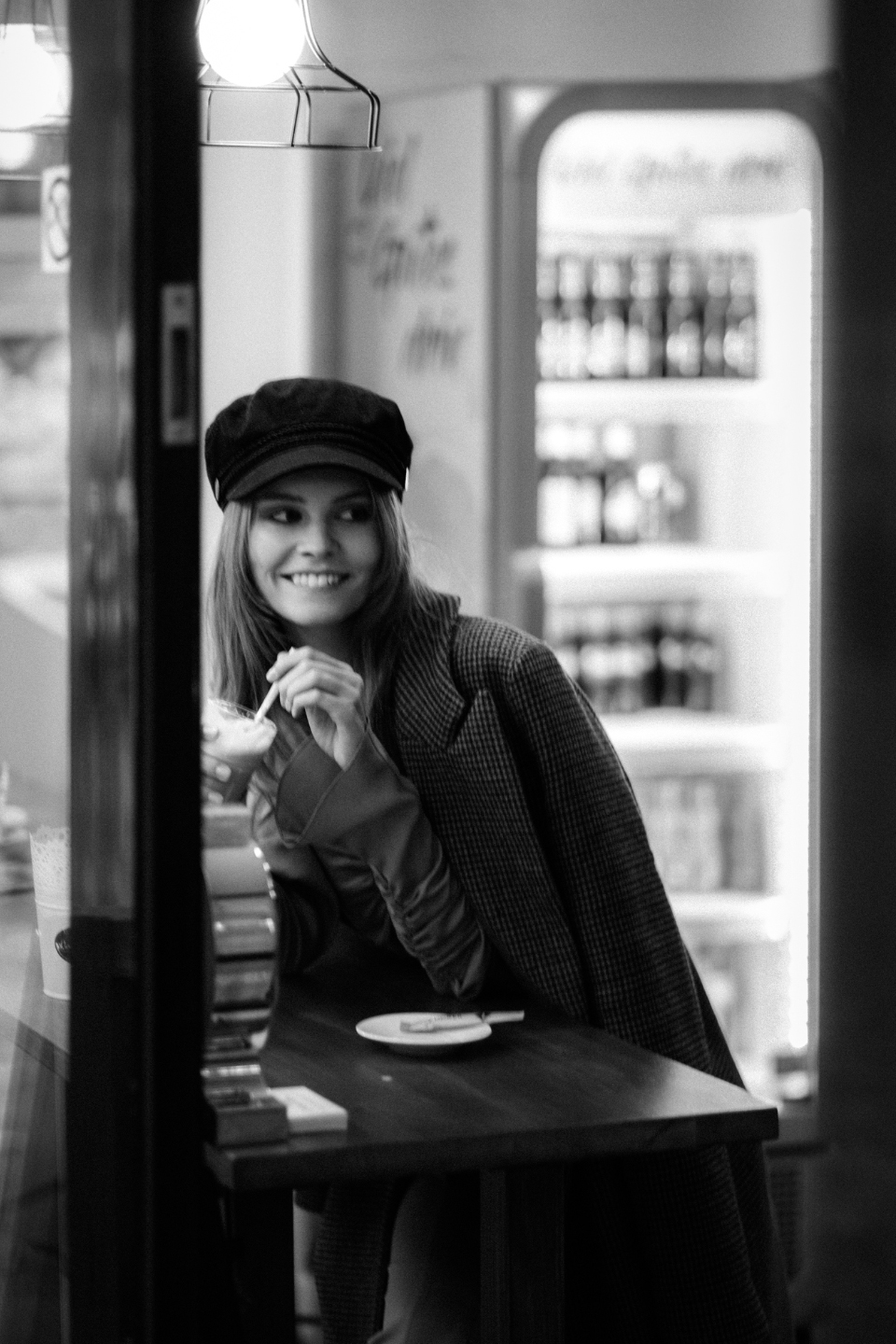 Schwarz-Weiß Portrait einer lächelnden Frau mit langen blonden Haaren und einem schwarzen Hut, die in einem Café sitzt und einen Kaffee trinkt.