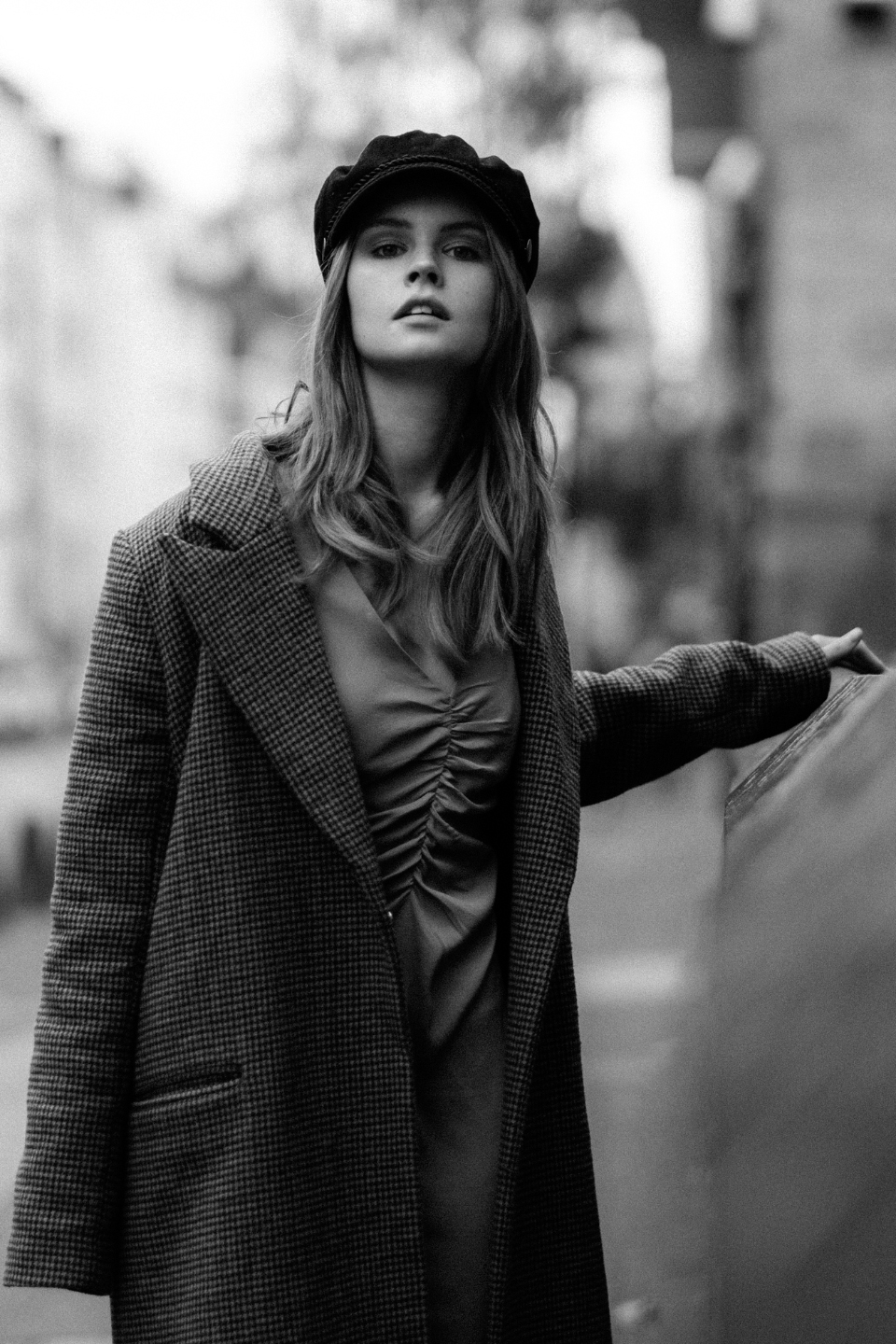 Schwarz-Weiß Portait einer Frau mit langen blonden Haaren, schwarzem Hut und kariertem Mantel, die durch die Straßen schlendert und dabei in die Kamera schaut