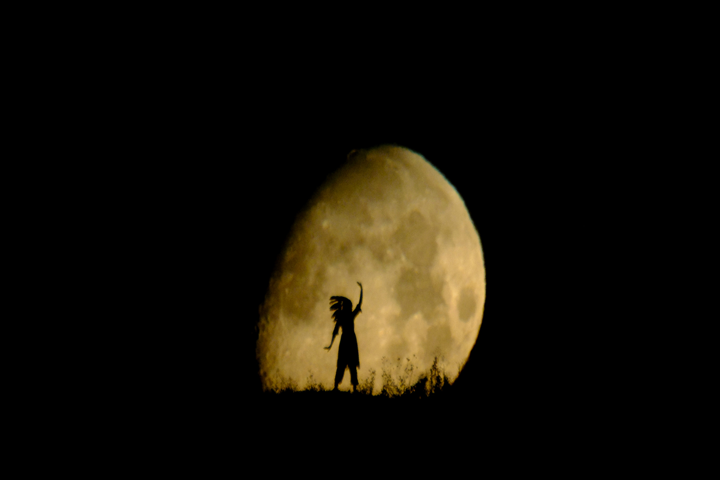 Nachtaufnahme des Mondes mit der dunklen Silhouette einer Person mit indianischem Federkopfschmuck im Vordergrund