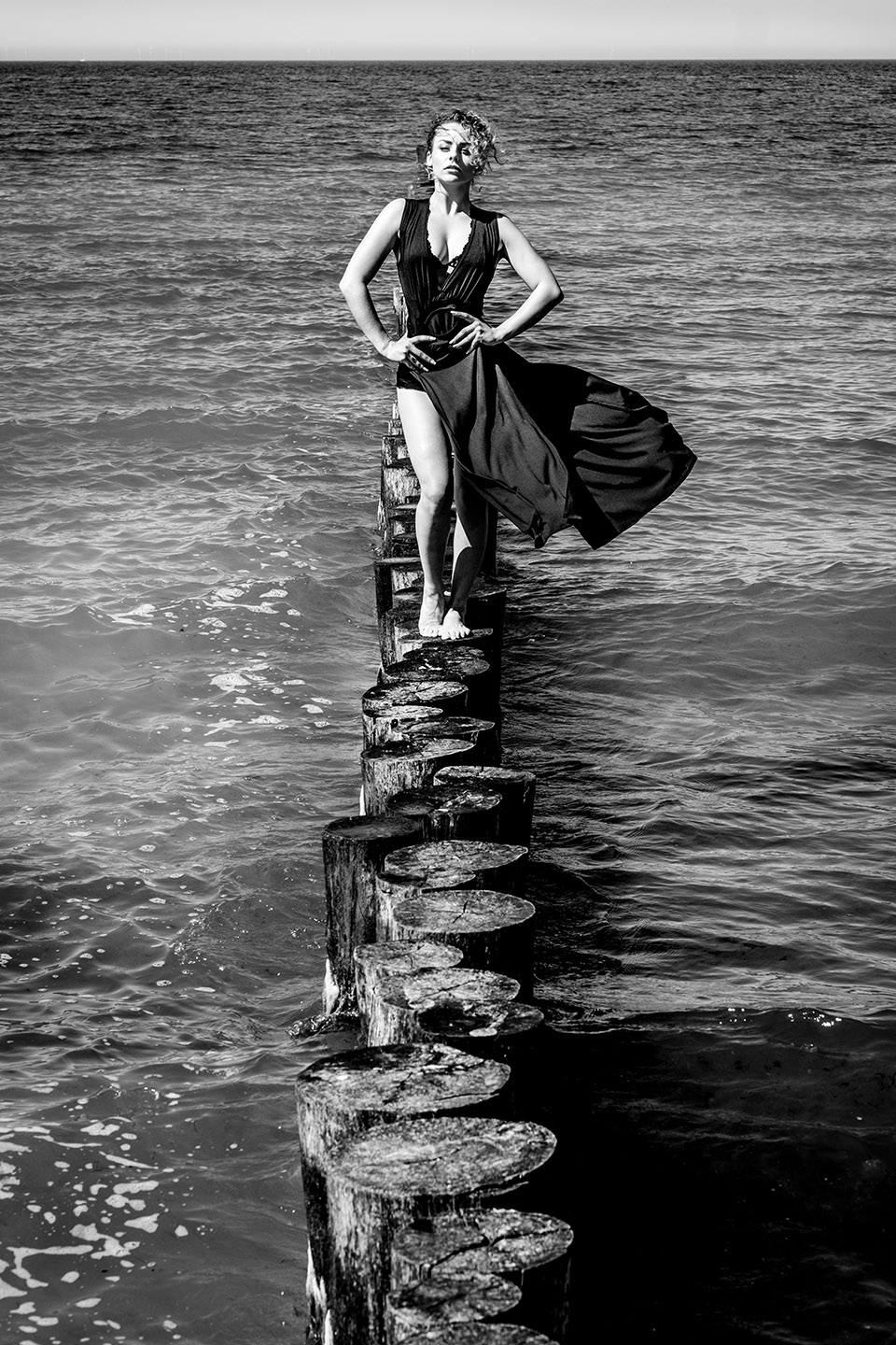 Schwarz-weiß Aufnahme einer Frau mit blonden Locken und langem schwarzen Kleid, die auf Wellenbrechern im Meer in Richtung Kamera läuft