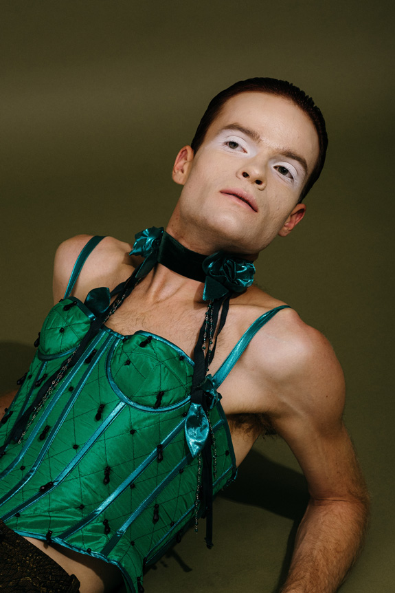 Portrait of man reclining back onto his elbows wearing eye shadow and a corset. Image taken in a studio environment