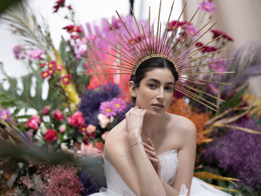 Portrait of a bride with a colourful head dress pictured in front of a vibrant floral display.