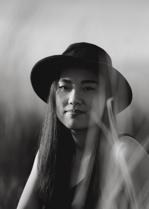 Black-and-white mid-frame portrait of young woman in a black wide-brimmed hat among long grass