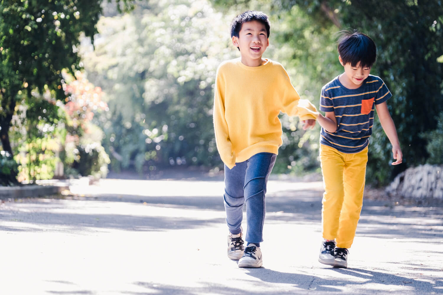 FUJIFILM Exposure Center Article - Be Versatile With Your Vision Using Zoom Lenses. Photograph shows a long shot of two boys walking towards the camera along a tree-lined driveway, holding hands and laughing.