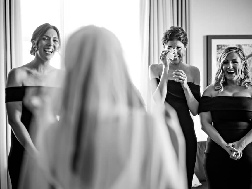 Professional photographer, Alison Conklin, on why the FUJIFILM GFX system is so good for location portrait photography. Image taken from behind a bride as she gets ready for her wedding standing in front of her bridesmaids who are positioned in front of a window.