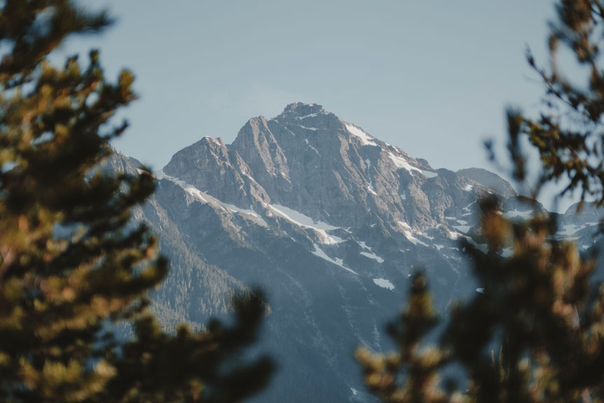 FUJIFILM Creators talk about how a lend can affect your message. Mio Monasch uses a mid-telephoto prime lens to frame a distend mountain, framed by pine trees in the foreground that are blurred.