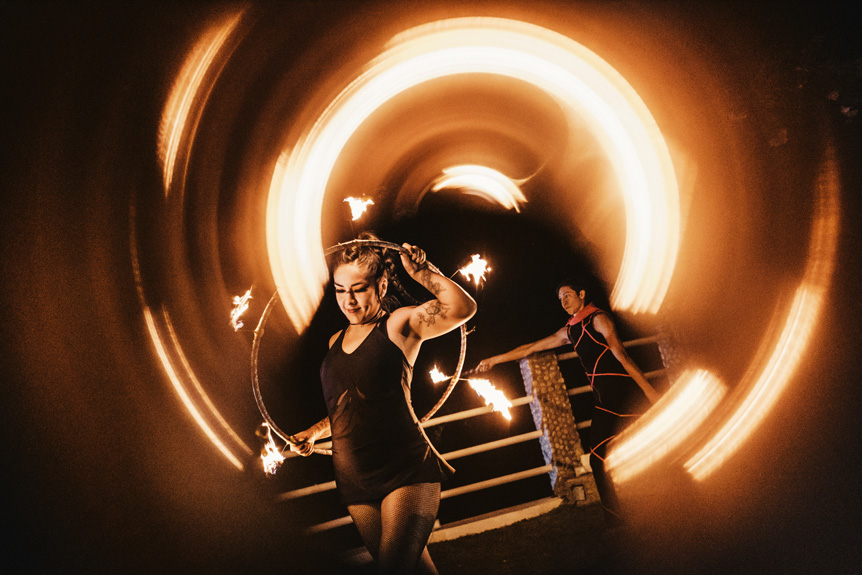 A young fire dancer poses with her flaming hoop while streaks of fire fly overhead