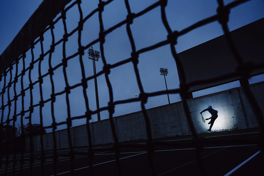 A tennis player serves at dusk, as seen through the gaps in the tennis net