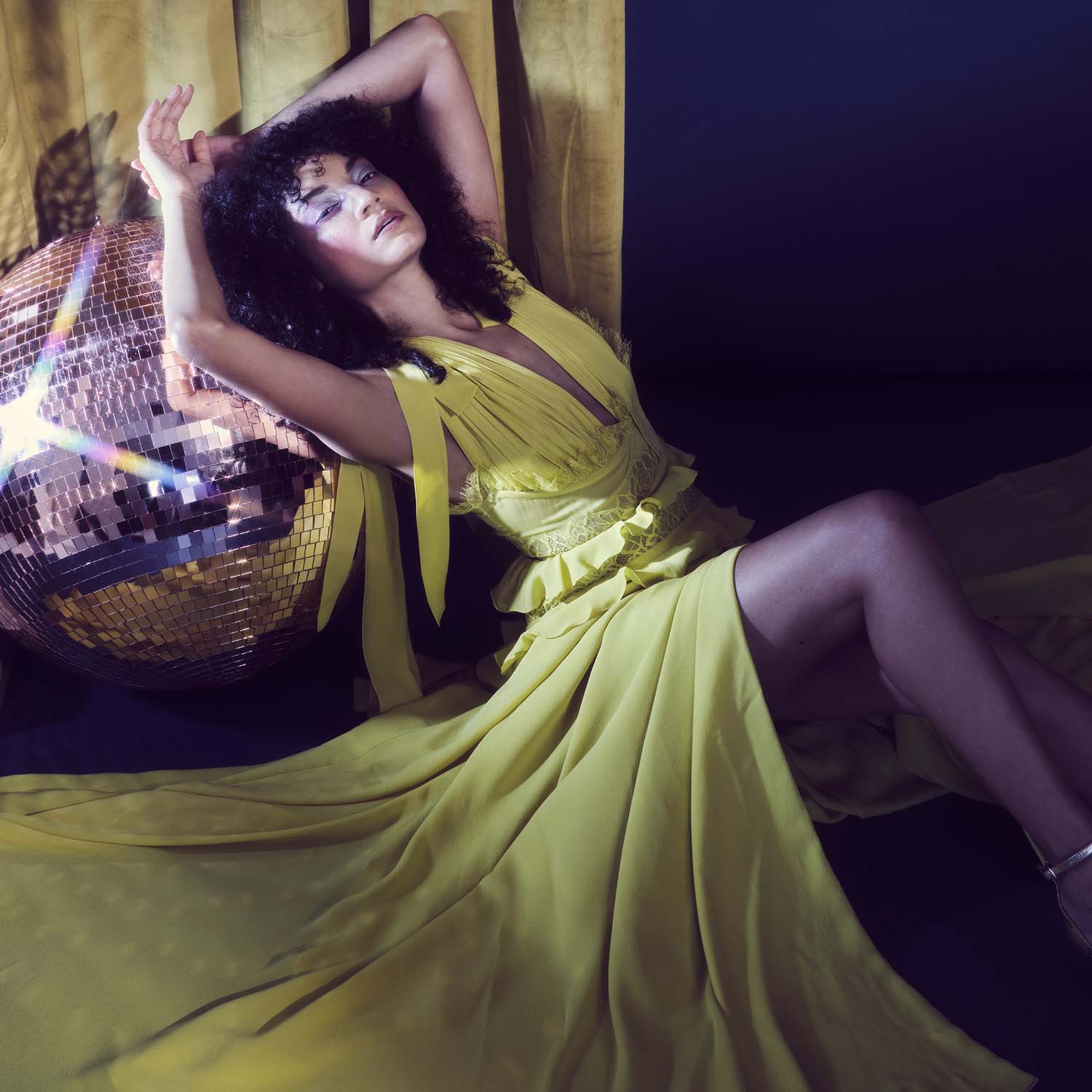 Model with dark, curly hair in flowing, yellow dress leaning against pink glitterball