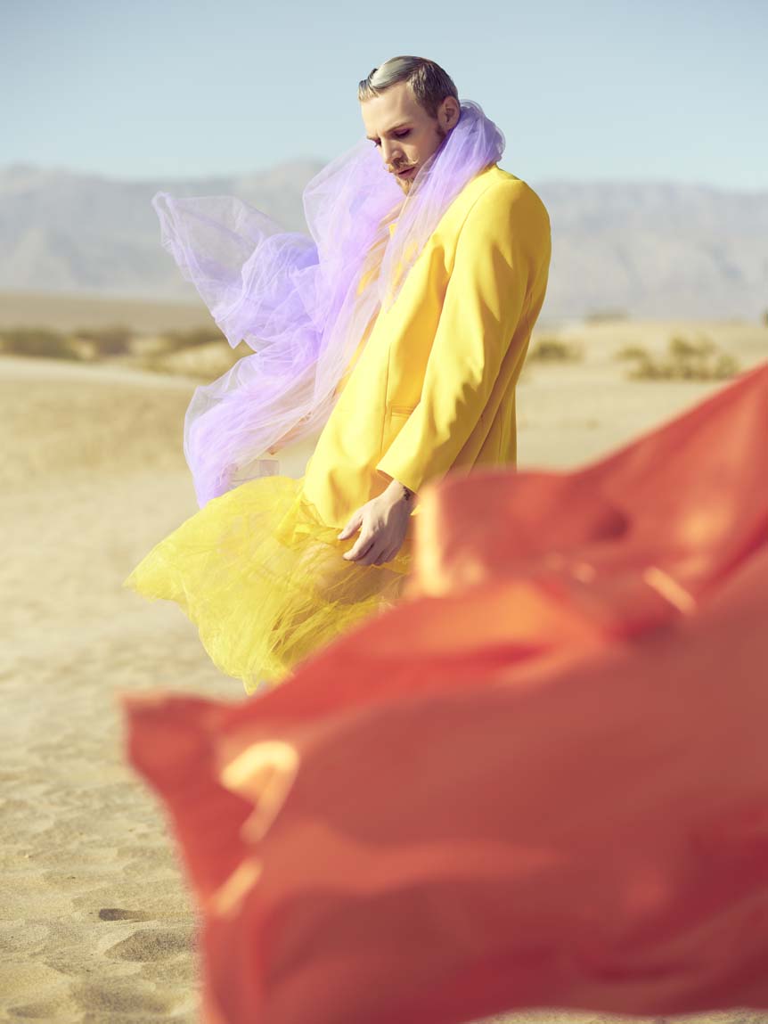 A man in a yellow chiffron suit-dress sways orange and pink scarves in the winds of Death Valley