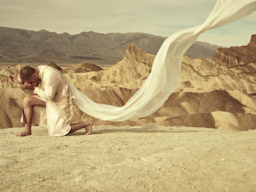 Clad in a flowing white gown, a man with a shaved head clutches his head in the spiritual sands of Death Valley