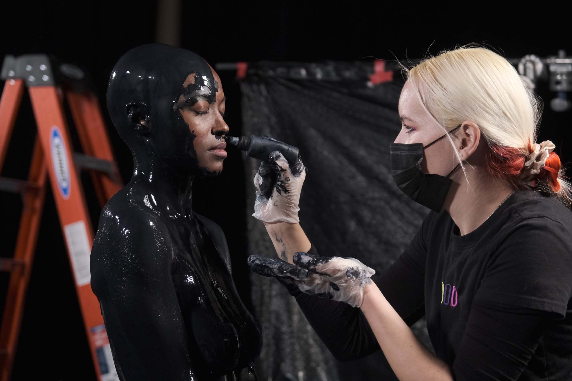 Soaked in black paint, a female model iis tended to by a make up artist