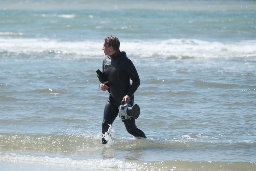 Filmmaker in wetsuit walking out of ocean with camera