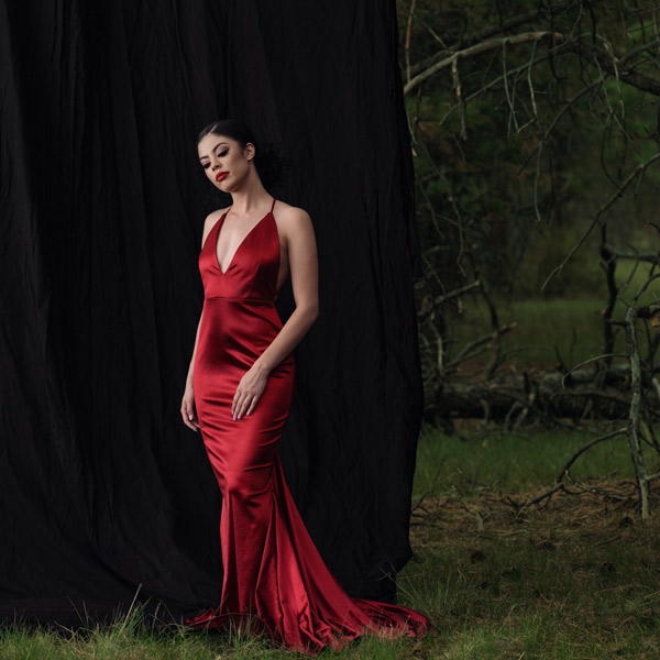 Beautiful woman in red dress, standing in woodland with black backdrop behind her