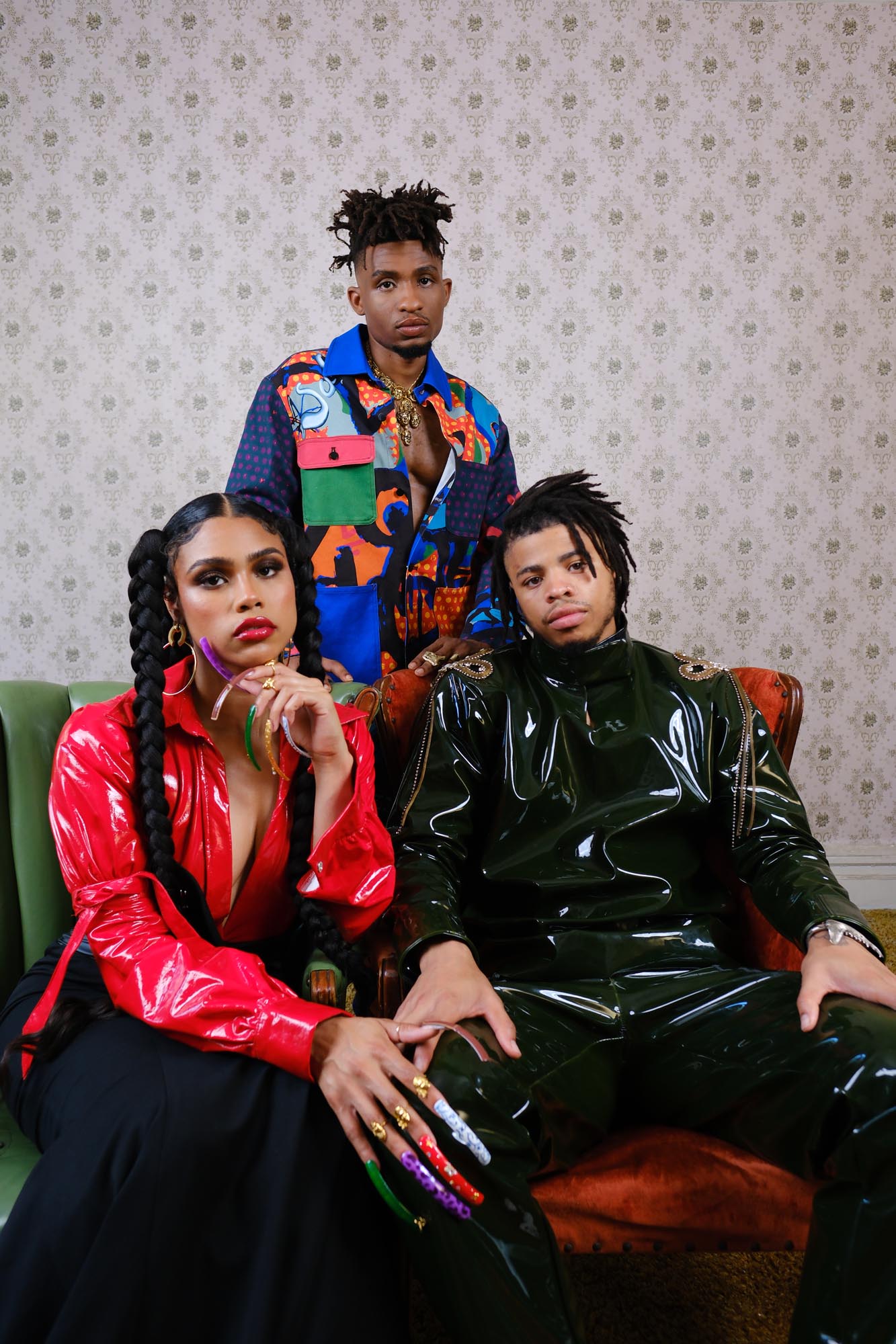 Three young African Americans pose in luxurious high fashion, framed against a drab wallpaper background