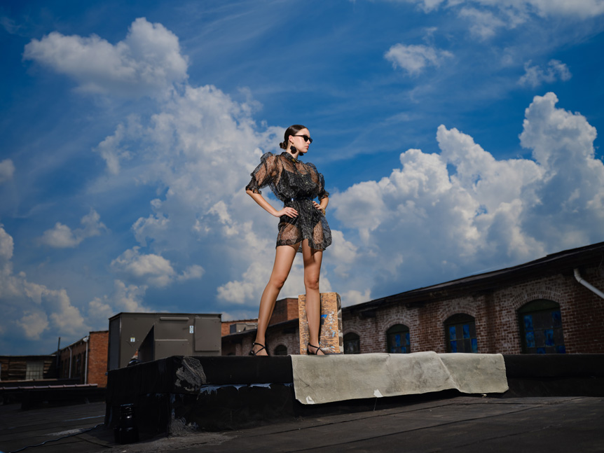 Young woman fashion model being photographed on rooftop under blue cloudy sky