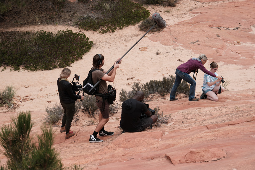 Photographer guiding young mentee in desert landscape with crew filming