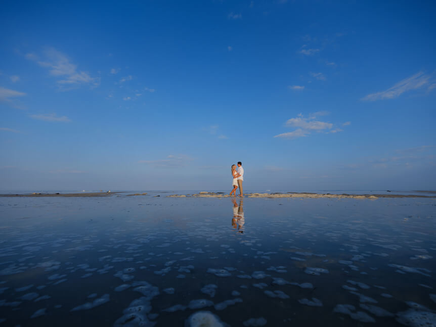 couple cuddling on beach. Wide shot to show expanse of blue sky and reflection in water