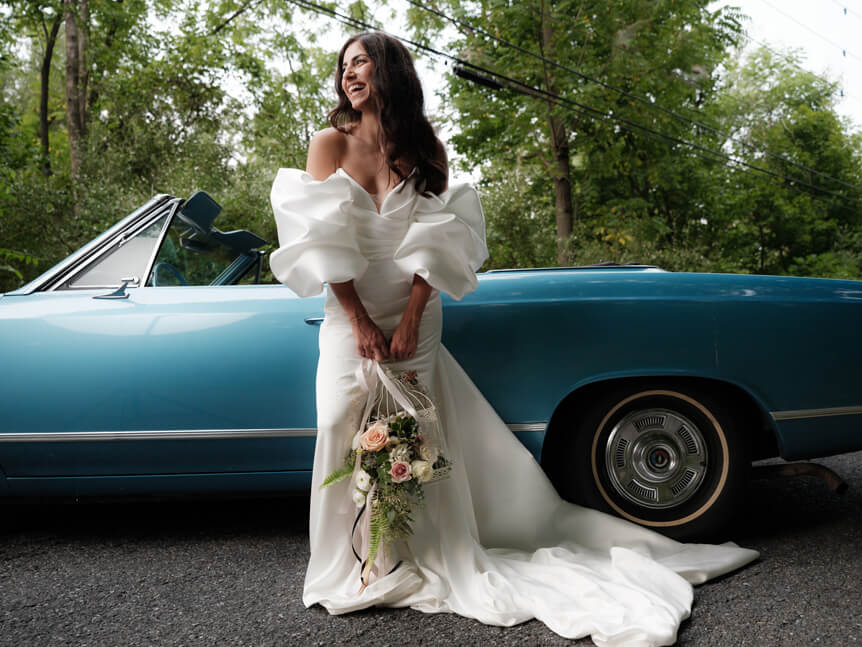 Bride with bouquet of flowers standing in front of blue classic American convertible car.