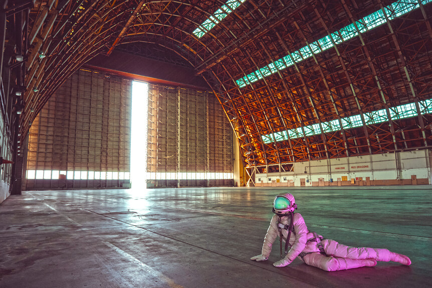 Astronaut reclining on concrete floor of abandoned blimp hangar.