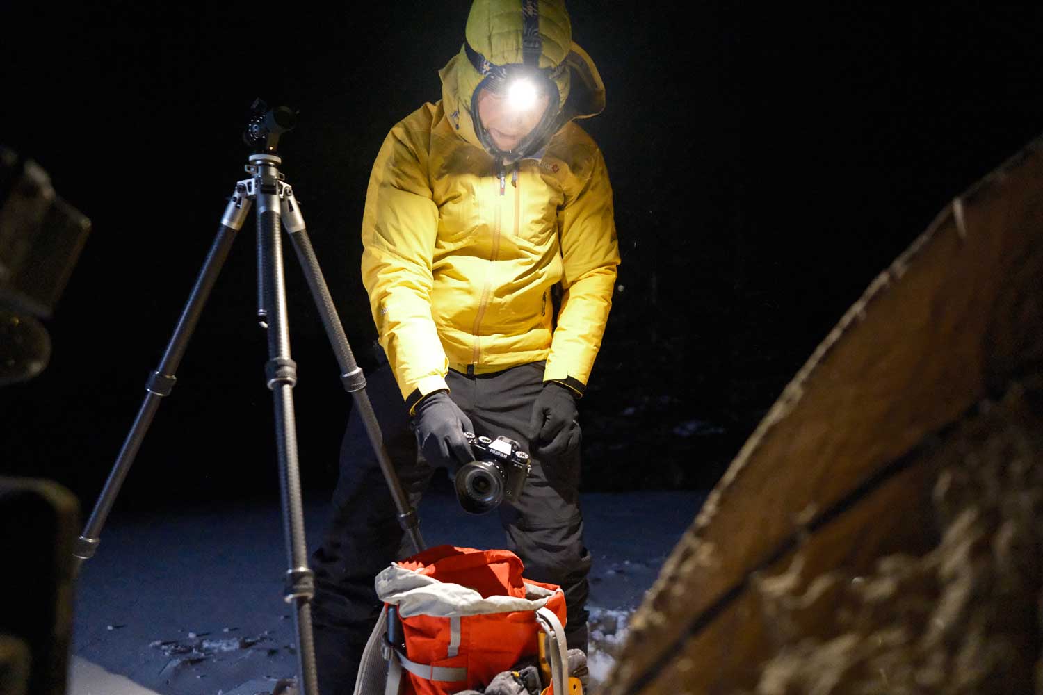 Seth K Hughes wearing yellow jacket and hood with head torch on holding a FUJIFILM X-T5 camera outside a tent next to a tripod in the snow at night