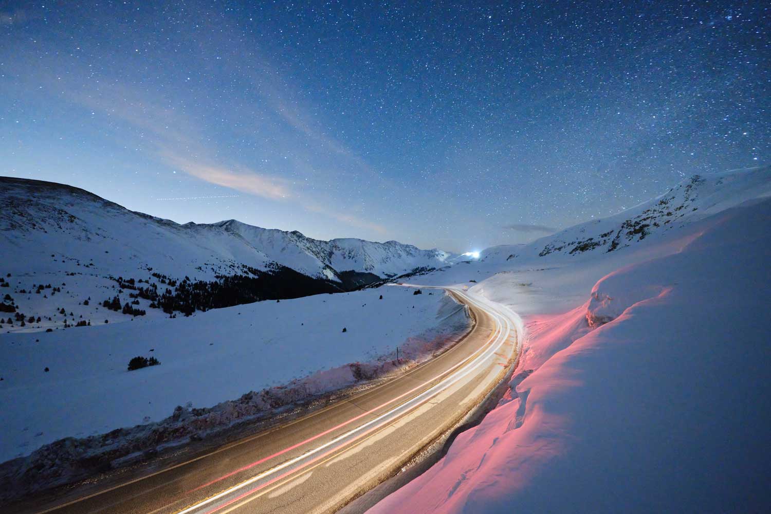 Astrophotography image of snowy valley at night below starry sky, with traffic trails winding along road in foreground and into the distance.
