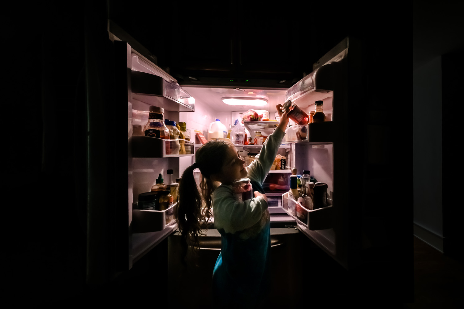Girl takin jar from fridge in dark room. Only illumination is coming from the refrigerator's interior light