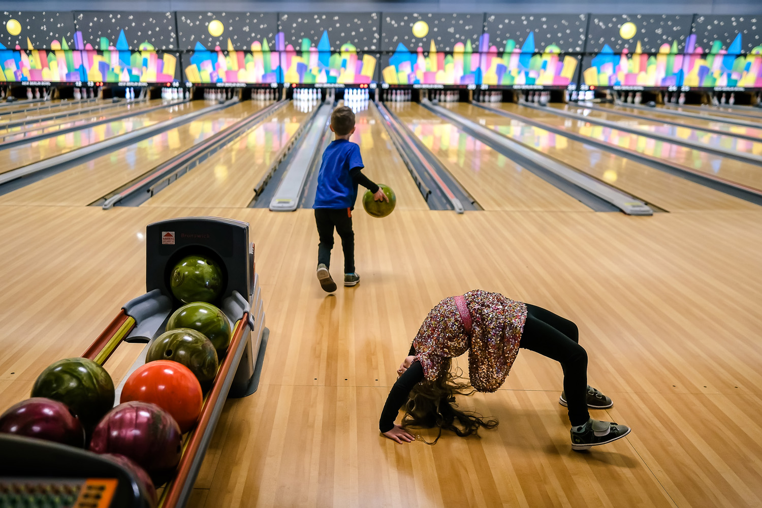 In a bowling alley a young girl performs 'the crab' gymnastics move, while a young boy bowls in the background