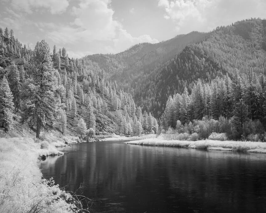 Monochrome infrared image of tree-lined river leading to mountain