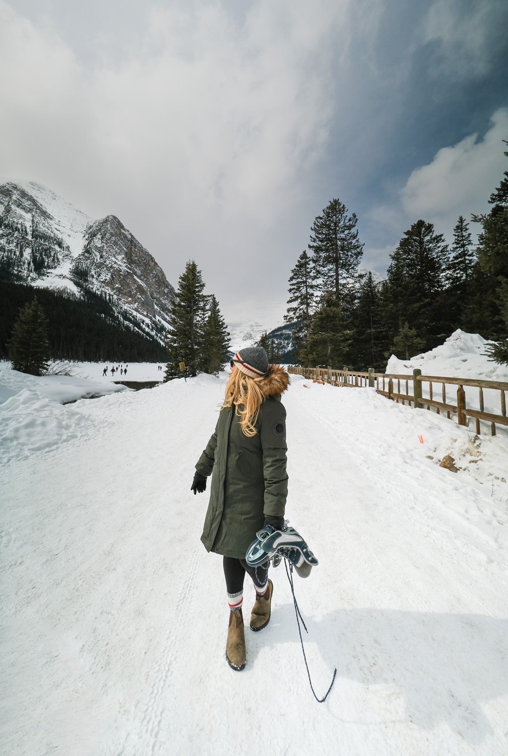 Girl in winter coat walks towards camera on snowy track with trees and mountains in background