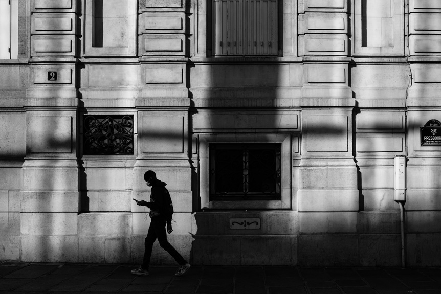 Side-on street photograph of silhouetted man looking at his phone while walking along pavement past a stone-fronted building