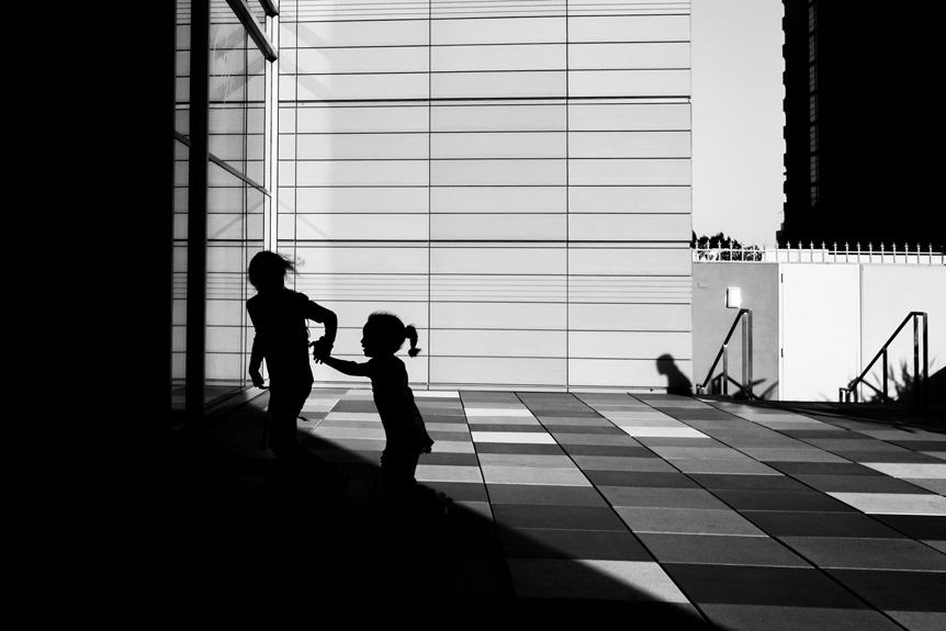 black and white image of a city square in bright sunlight with the silhouettes of two children holding hands in the foreground.