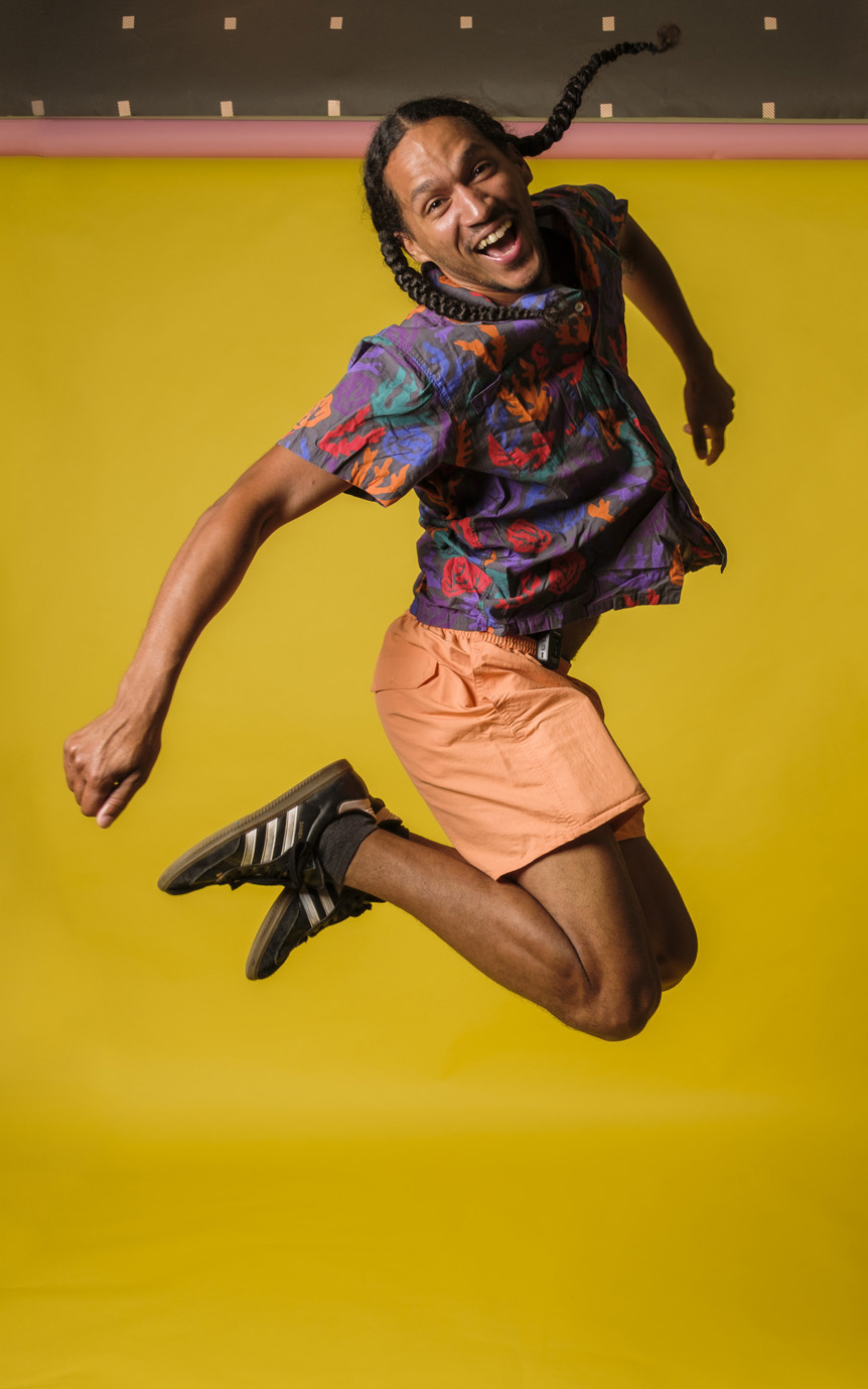 Young man with long braids jubilantly jumping into air against yellow backdrop