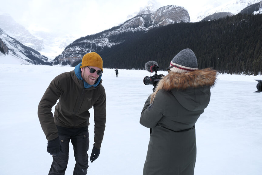 Woman filming man standing on frozen lake with mountains in background