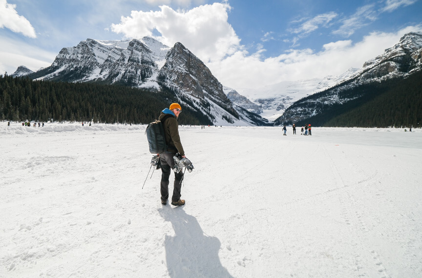 Man hiking across open snowy ground looking over his shoulder back at the camera. There are snowy mountains in the background