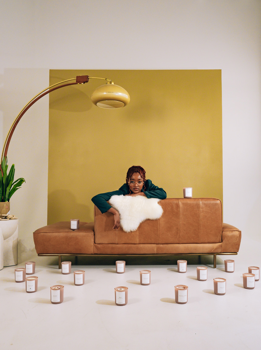 Candle business owner leans over the back of a brown leather sofa with yellow background in 'Green Spaces' hub. Candles surround her on the floor.