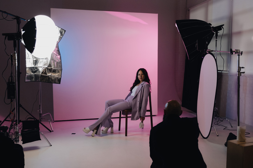 Wide shot of a photography studio where a girl with brown hair is wearing a pink suit posing for a photograph. There are lights and a camera pointed in her direction