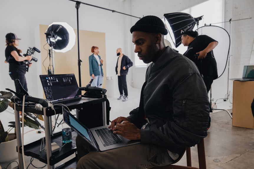 Behind the scenes of a photoshoot, depicting the process. A man on a laptop is in the foreground while the photoshoot happens behind him